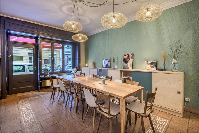 Der Raum der Concept Kitchen mit Holztafel, Stühlen, Fliesenboden und runden Hängeleuchten